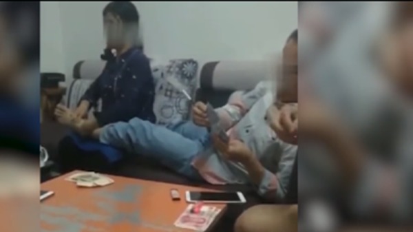 視頻中，一名男子半躺在沙發上，一邊抽煙打牌，一邊把讓旁邊一女子替他捏腳。（視頻截圖）