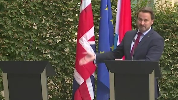 一脸不满的卢森堡总理贝特独自站在台上，朝着身旁英国国旗前空荡荡的讲台，大肆奚落着缺席的英国首相强生。